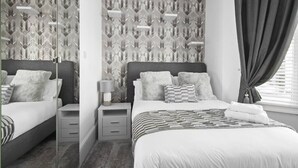 A modern bedroom with a patterned wallpaper, a bed with multiple pillows, a bedside table with a lamp, and a window with curtains. - The Roost Group - 1 Bedroom - Ground (Gravesend)