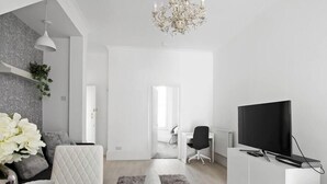 A modern living room with a television, a white sofa, a desk, and a chandelier. - The Roost Group - 1 Bedroom - Ground (Gravesend)