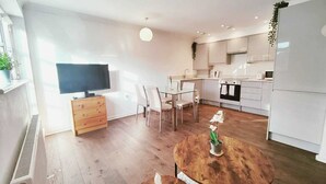 Una cocina moderna con armarios blancos, un comedor con mesa y sillas de madera, y un televisor a la izquierda.