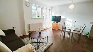 Una sala moderna con un sofá, una mesa redonda de centro con una planta, una zona de comedor con una mesa de vidrio y sillas, y un televisor al fondo.