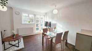 Una sala de estar moderna con una mesa de comedor de vidrio, sillas cómodas, un ventanal con persianas, un radiador y un televisor.