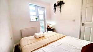 Un dormitorio con cama, ventana, radiador, mesita de noche con lámpara y puerta.