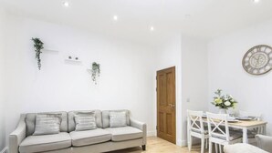 A modern living room with a sofa, a dining table, and chairs. There is a wooden door and a wall-mounted clock.