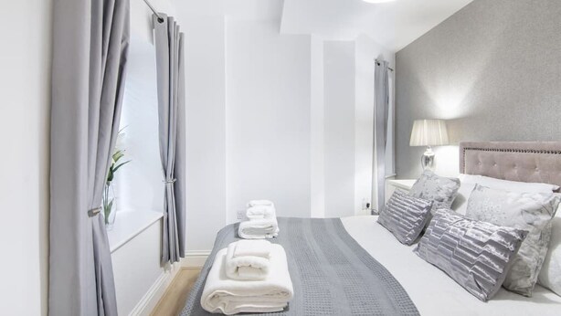 Habitación de hotel con cama, mesita de noche, lámpara, ventana con cortinas y un perchero para toallas.