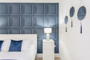 A bedroom with a white bed, blue pillows, a white nightstand, a blue wall panel, and two wall-mounted mirrors.