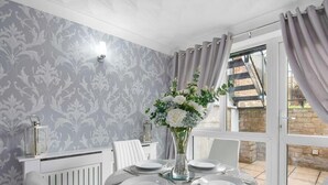 Un coin repas avec une table dressée pour quatre personnes, un vase rempli de fleurs, un radiateur et une fenêtre avec des rideaux.