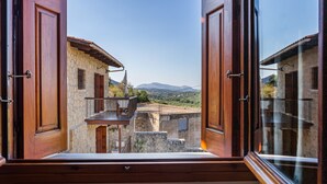 A view from a window with open shutters, revealing a stone building with a balcony and a mountainous landscape in the background. - Olive Villas (Platanias)