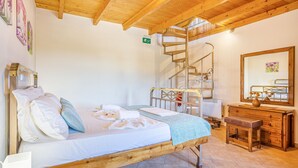 A bedroom with a wooden bed, a small bench, a dresser, a mirror, and a spiral staircase. - Olive Villas (Platanias)