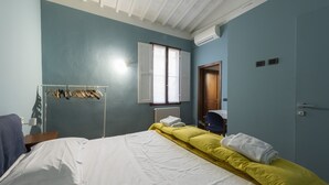 A bedroom with a bed, a window, an air conditioning unit, a door, and a clothing rack. - Toscanella 3 Bedrooms by Mmega (Florence)