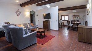 A spacious living area with a sofa, armchair, and coffee table, wooden beams on the ceiling, a television, and a dining area with chairs and a table. - Toscanella 3 Bedrooms by Mmega (Florence)