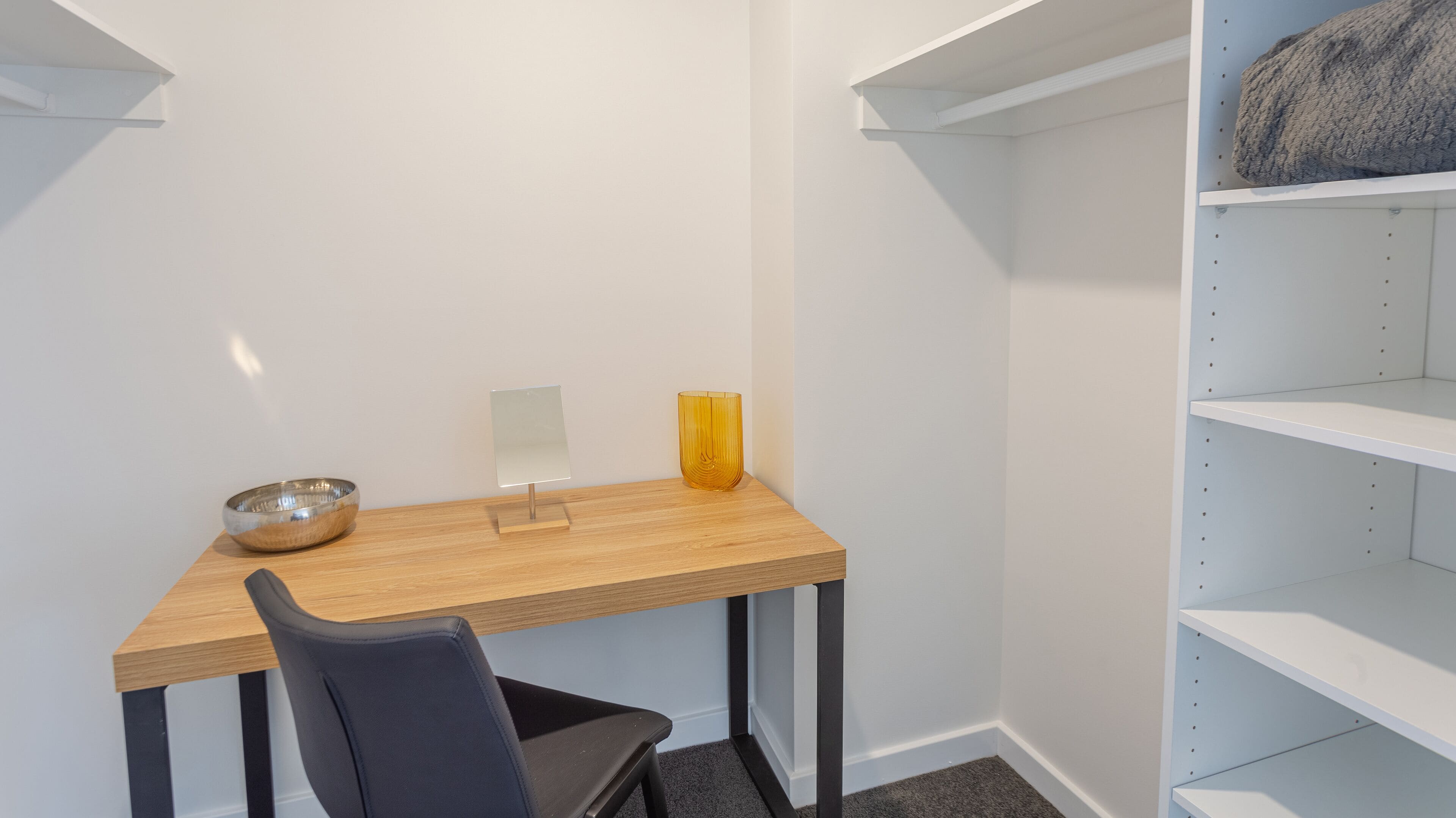 A small, modern room with a wooden desk, a chair, a small table with a bowl, a yellow vase, and white shelving units.