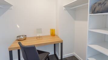 A small, modern room with a wooden desk, a chair, a small table with a bowl, a yellow vase, and white shelving units.