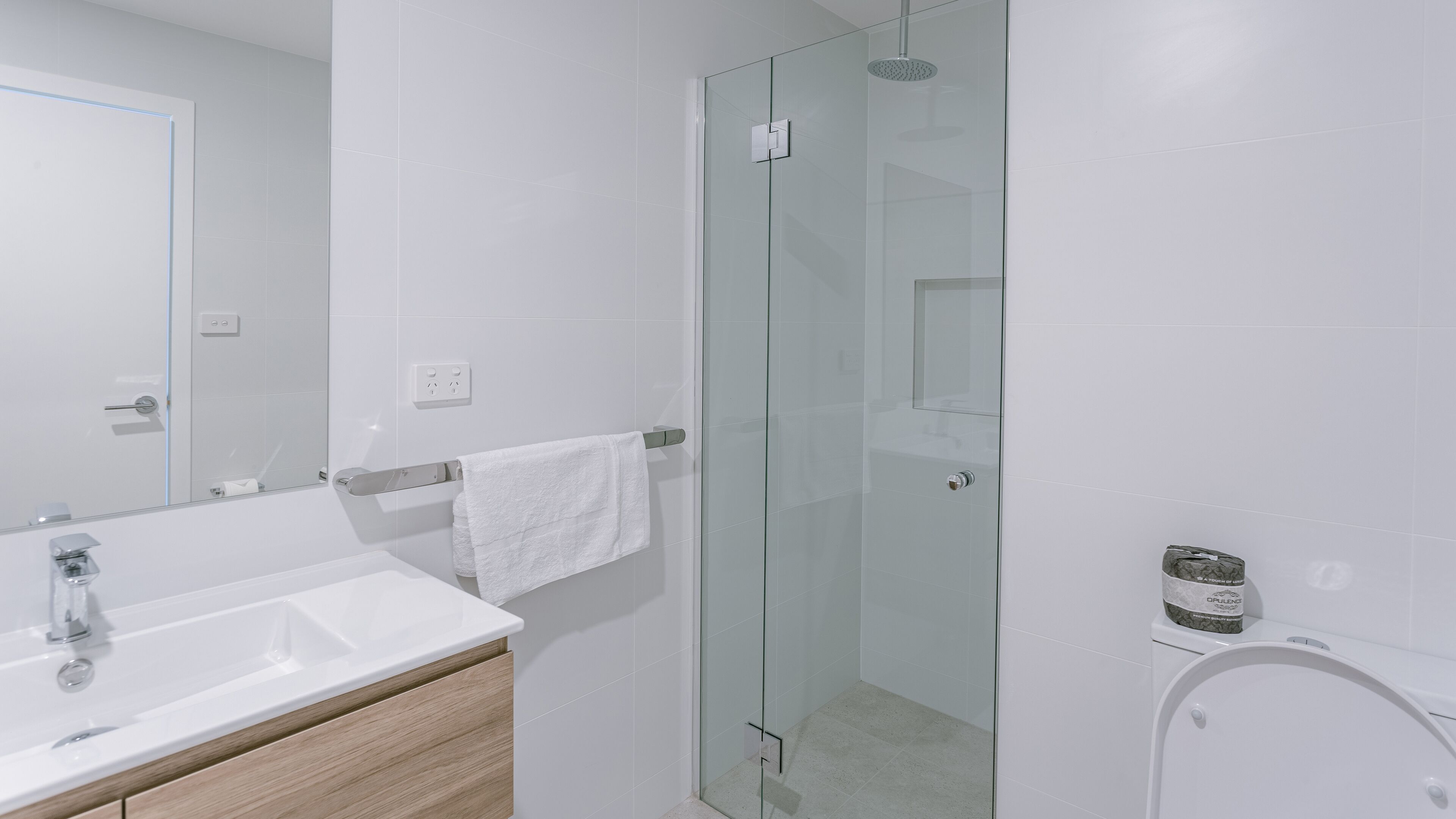 Un baño moderno con cabina de ducha de vidrio, un lavamanos blanco sobre un mueble de madera y un inodoro blanco.
