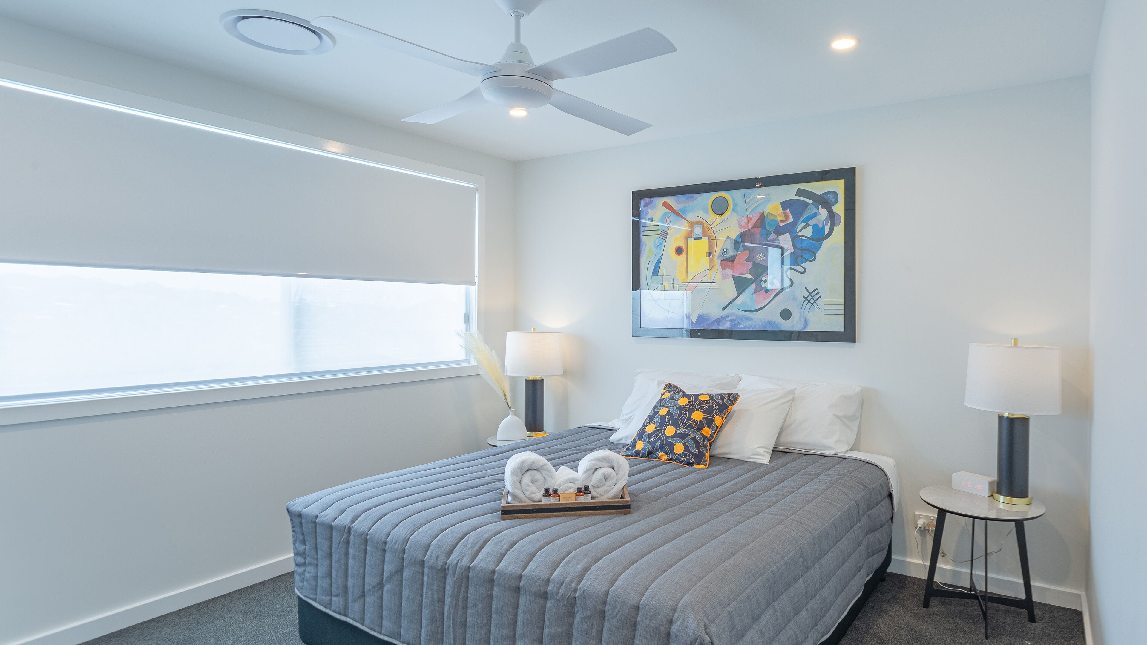 A modern hotel room with a large bed, two bedside lamps, a ceiling fan, a window with blinds, and a colorful abstract painting on the wall.