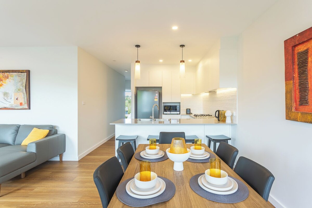 A modern living room with a grey sofa, a dining table set with white plates and bowls, and a kitchen in the background with a microwave and a coffee maker.