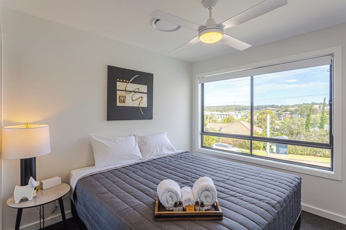 A hotel room with a large bed, a ceiling fan, a window with a city view, and a bedside table with a lamp.