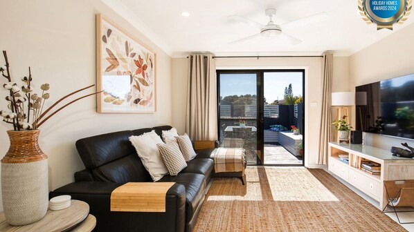 Un salón moderno con un sofá de cuero negro, una mesa de centro de madera, un televisor de pantalla plana grande y vistas a un balcón con mobiliario exterior.