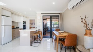 A modern kitchen with white cabinetry, a central island, and a dining area with a wooden table and chairs. - Alkira - Pet Friendly - 4 Mins Walk to Beach (Culburra Beach)