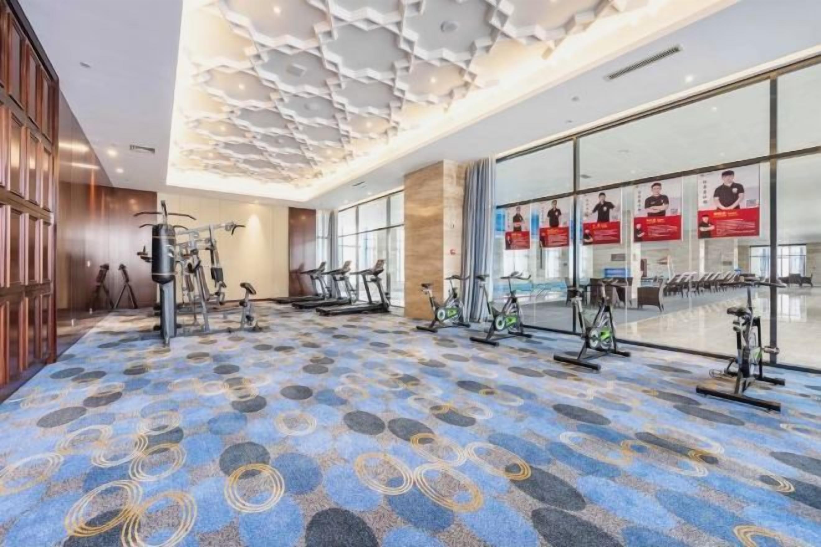 A hotel gym with exercise equipment, patterned carpet, and large windows.