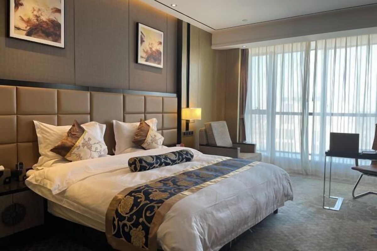 Hotelkamer met een groot bed, een bureau en een stoel. Er is een raam met gordijnen en kunstwerken aan de muren.