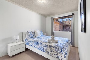 A bedroom with a bed, a nightstand, a window with a view of a brick building, and a ceiling fan. - Breakers 6 - Ocean Views at The Breakers (Forster)
