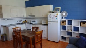 Cocina compacta con armarios blancos, microondas, estufa y refrigerador. Hay una mesa de comedor con cuatro sillas y un pequeño basurero. Un ventilador blanco está sobre una repisa, y se ve una pared azul de fondo.