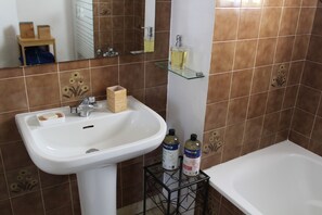 Une salle de bain avec un lavabo blanc, une étagère en verre où sont rangés les articles de toilette et une baignoire.