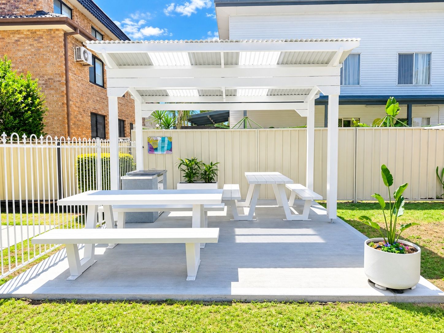 A covered patio area with white tables and benches, a potted plant, and a white fence.