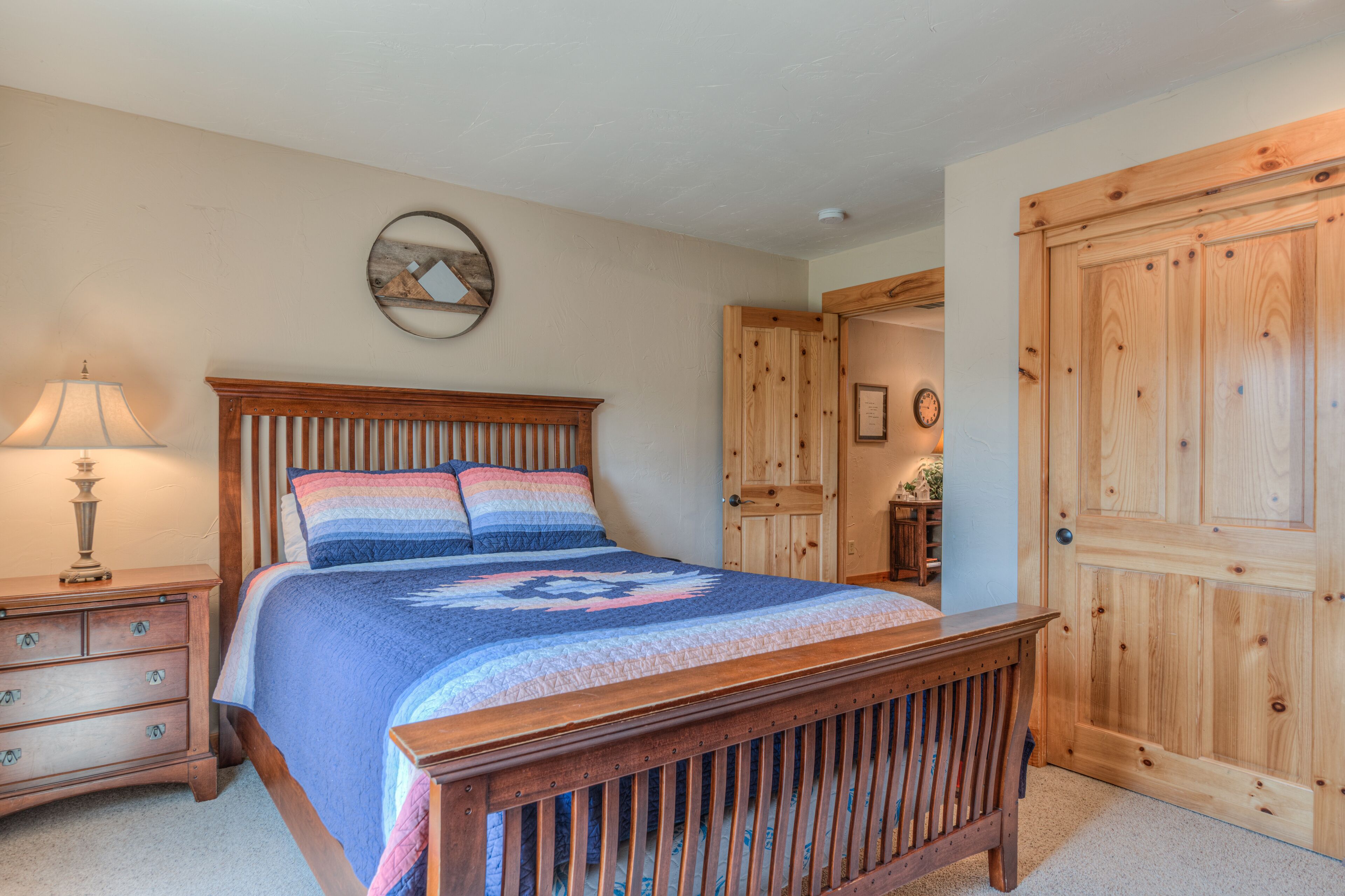 A bedroom with a wooden bed, a nightstand, a lamp, a wall clock, and a wooden door.