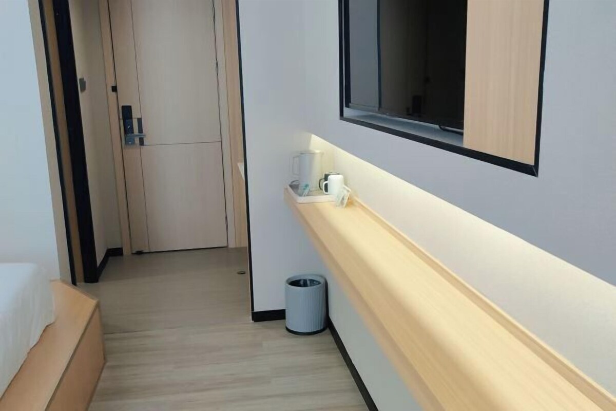A hotel room with a bed, a small bedside table, a trash bin, a wall-mounted shelf with a coffee maker, and a window.