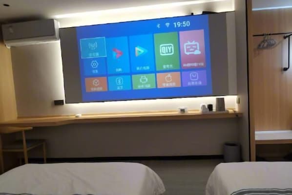 A hotel room with a large screen displaying a mobile interface, two beds, a desk, and a wardrobe.