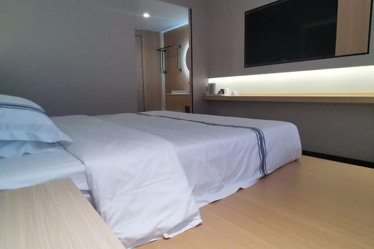 A modern hotel room with a neatly made bed, a flat-screen TV mounted on the wall, and a small bedside table with a lamp.