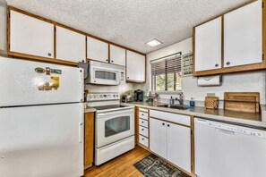 A compact kitchen with white appliances, wooden cabinets, and a window with blinds.