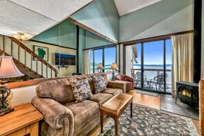 A cozy living room with a sofa, a coffee table, and a fireplace. There is a staircase leading to an upper level, and large windows offering a view of the ocean. - Beach Front Condo, Views, Views Views (South Lake Tahoe)