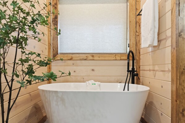 A bathroom with a freestanding bathtub, wooden walls, a window, and a plant.