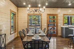 Comedor de madera con una larga mesa dispuesta para una comida, rodeada de sillas y una araña colgante.
