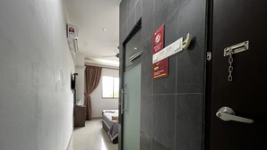 Quarto de hotel com cama, escrivaninha, cadeira, janela com cortinas, porta com placa e ar condicionado fixado na parede.