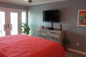 Une chambre avec un grand lit, un téléviseur fixé au mur, une commode en bois et une plante en pot.