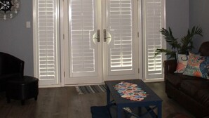 Uma sala de estar com ventilador de teto, relógio, porta com persianas, mesa de centro e sofá.