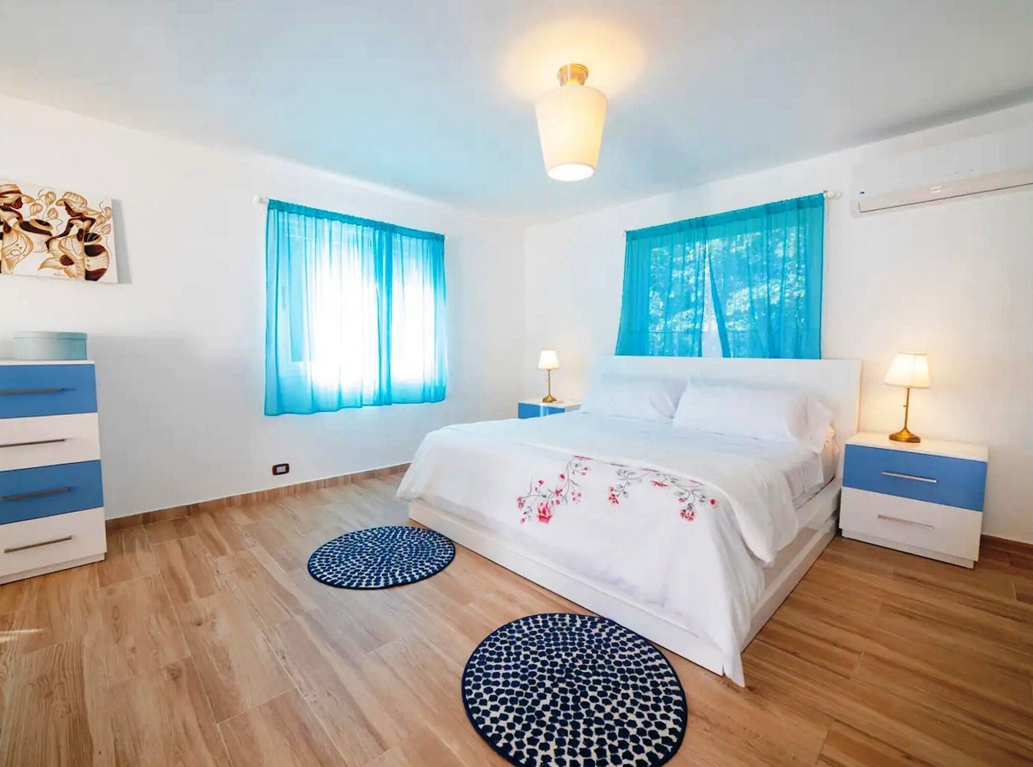 A bedroom with a bed, two bedside tables, a dresser, two windows with blue curtains, a ceiling light, and two round rugs.