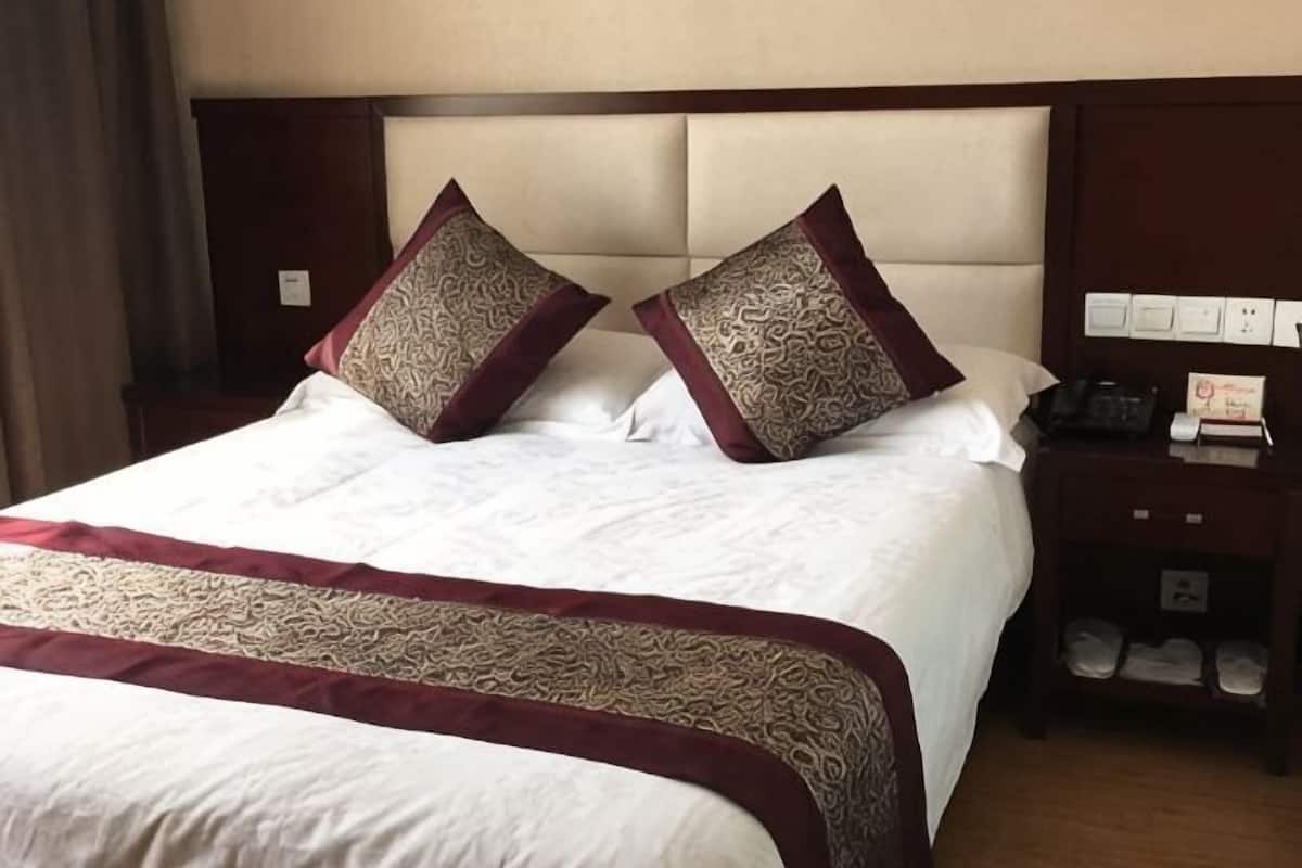 Hotellihuone, jossa on suuri sänky, kaksi koristeellista tyynyä, yöpöytä, jossa on puhelin ja lamppu, sekä puinen yöpöytä, jossa on kenkäteline.