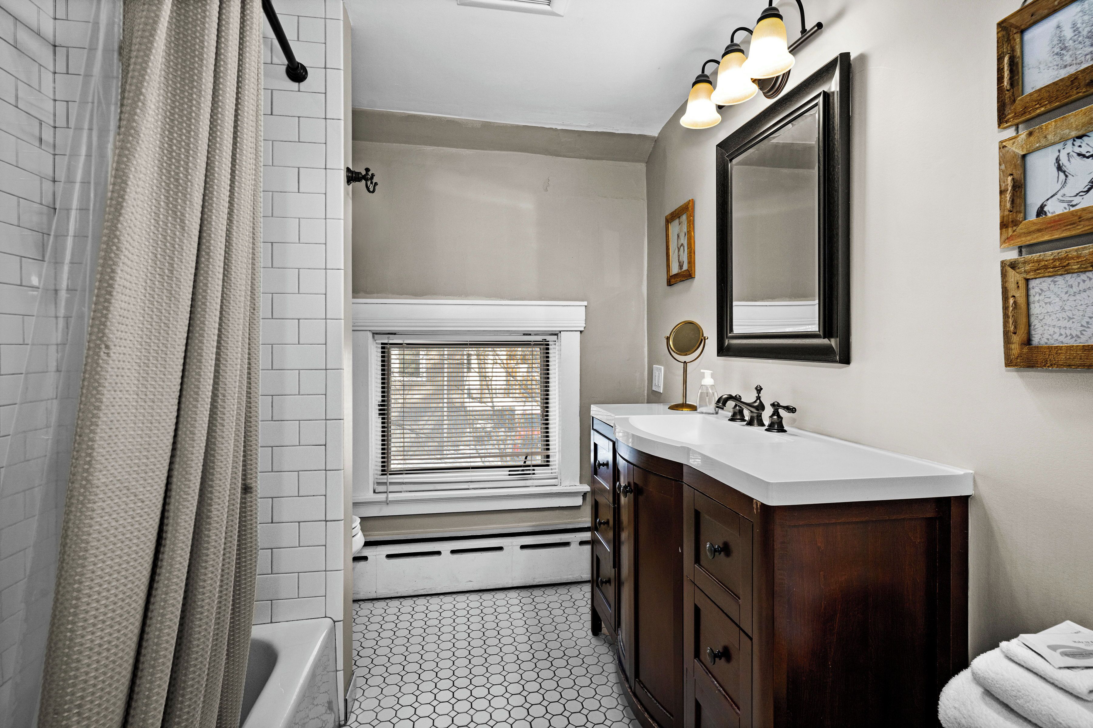 A bathroom with a bathtub, a window with blinds, a vanity with a mirror, and a tiled floor.