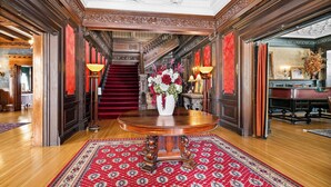 Un lobby espacioso con una escalera de madera, una mesa redonda de madera y un jarrón de flores, y una alfombra roja con diseño.
