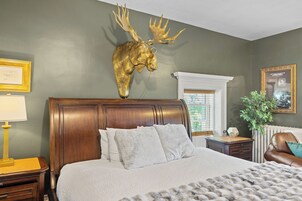 Un dormitorio con una cama grande, cabecera de madera, cabeza de venado montada, mesita de noche, lámpara, planta en maceta y un cuadro en la pared.