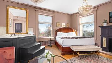 A bedroom with a large bed, a vanity with a mirror, a red armchair, and a chandelier.