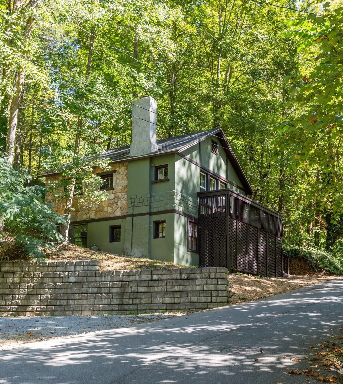 Una casa a due piani con facciata in pietra, un camino e un portico recintato, immersa in alberi fitti.