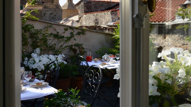 A view through a window of a dining area with a table set for tea, pastries, and flowers. A bell tower is visible in the background. - La Terrazza di Vico Olivi B&B  (Ventimiglia)