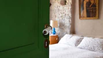 Une porte verte avec un trou de serrure et une serrure. Au fond, on aperçoit un lit avec une literie blanche et une lampe sur une table de chevet.