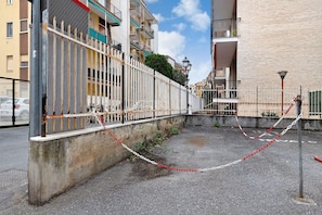 Une cour fermée par un portail et une chaîne, un mur en béton et, au fond, un immeuble doté de balcons.