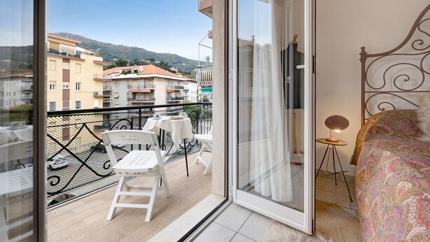 Ein Balkon mit Tisch und Stühlen, ein Bett mit gemustertem Überwurf und Blick auf Gebäude und Hügel.